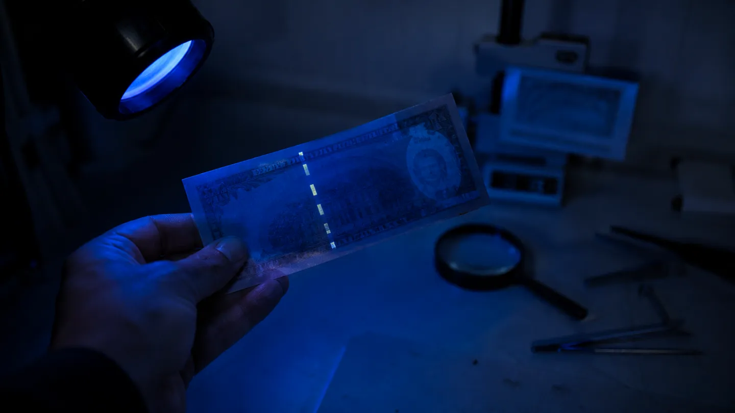 A hand examining a genuine U.S. banknote under UV light, highlighting security features relevant to detecting counterfeits.