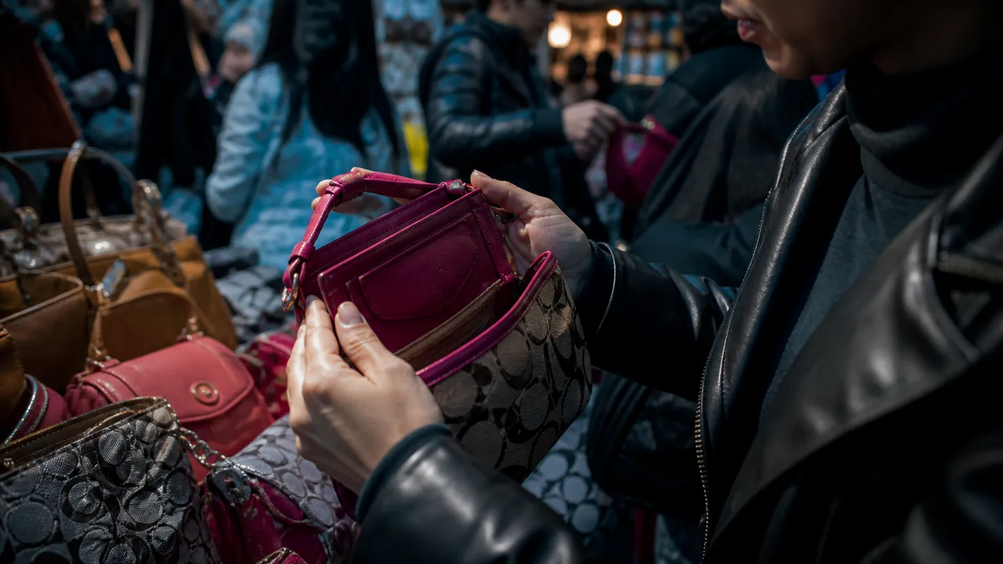 A person closely inspecting counterfeit designer handbags at a market stall, reflecting the article's focus on counterfeiting.